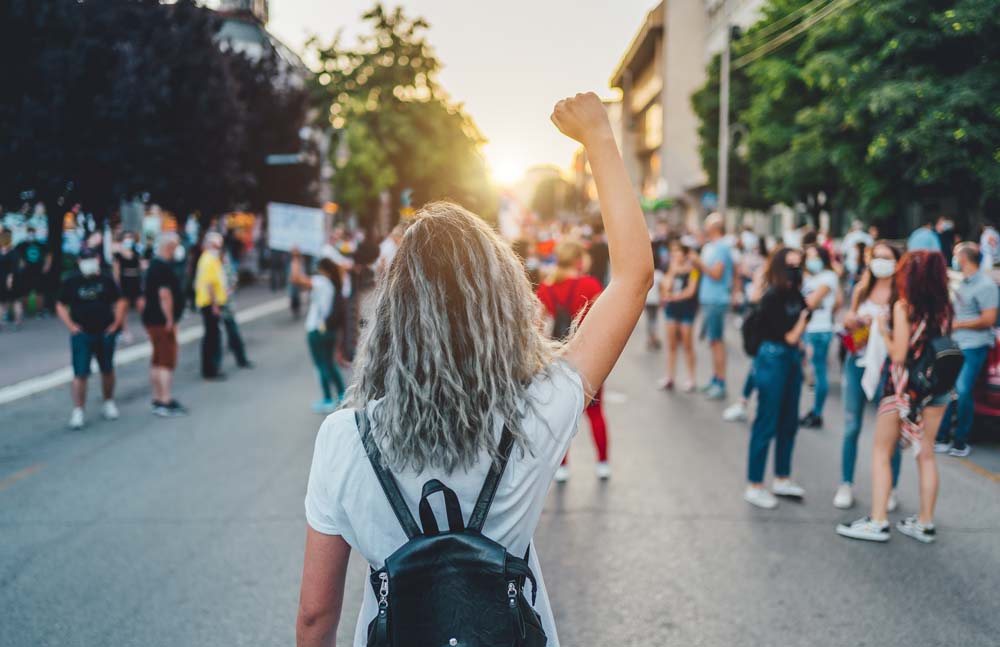 Woman facing away toward group of people protesting with her fist raised.